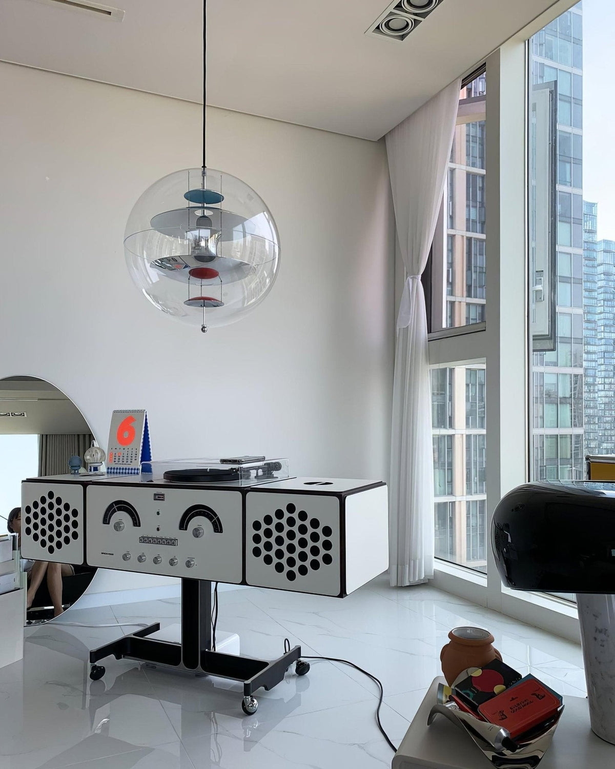 This Clérachic sphere pendant hangs in a white corner room with floor-to-ceiling windows, above a retro radio cabinet on a black stand.