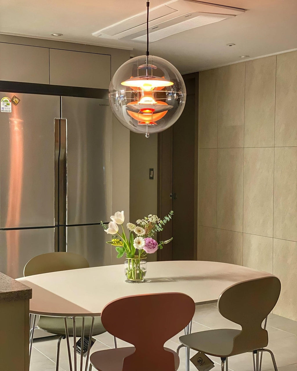 Glowing orange, this Clérachic sphere pendant hangs over a round table with brown and olive chairs, next to a steel fridge.