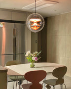 Glowing orange, this Clérachic sphere pendant hangs over a round table with brown and olive chairs, next to a steel fridge.