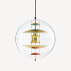 A Clérachic sphere pendant in gold on a plain backdrop, with brass discs, teal and coral dishes and a small brass weight beneath.
