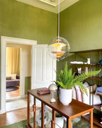 A lit Clérachic sphere pendant hangs in a green-walled room with a high ceiling, above a wooden sideboard, a fern and a small table lamp.