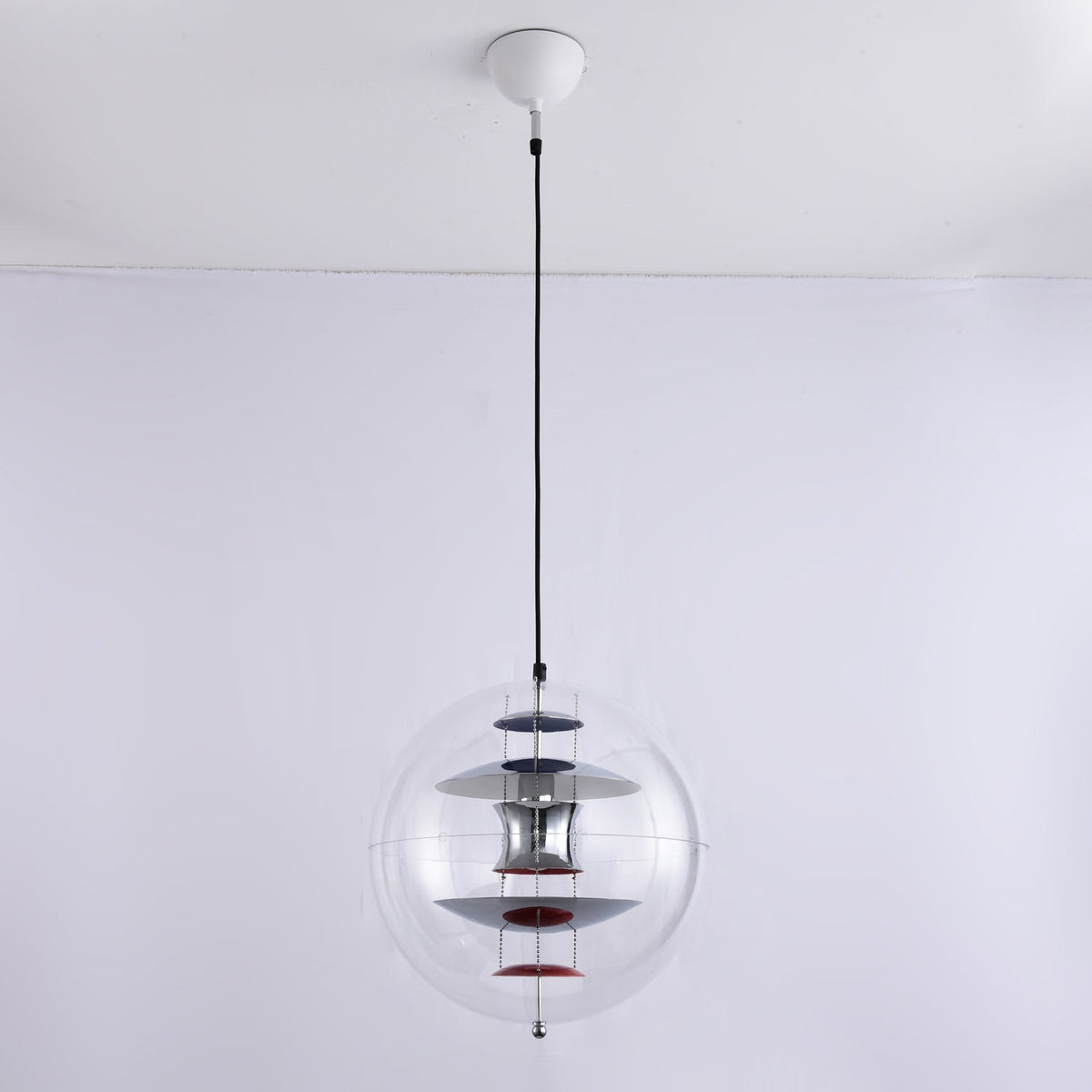 A Clérachic sphere pendant with a white ceiling canopy and black cord hangs against a pale wall, its chrome discs and red dish unlit.