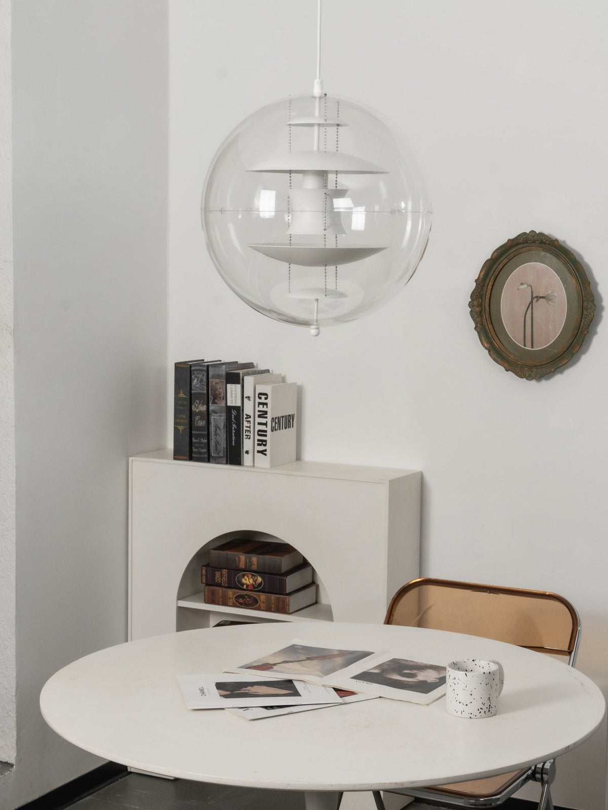 An unlit white Clérachic sphere pendant hangs above a white round table, with a shelf of books and a round olive-framed mirror.
