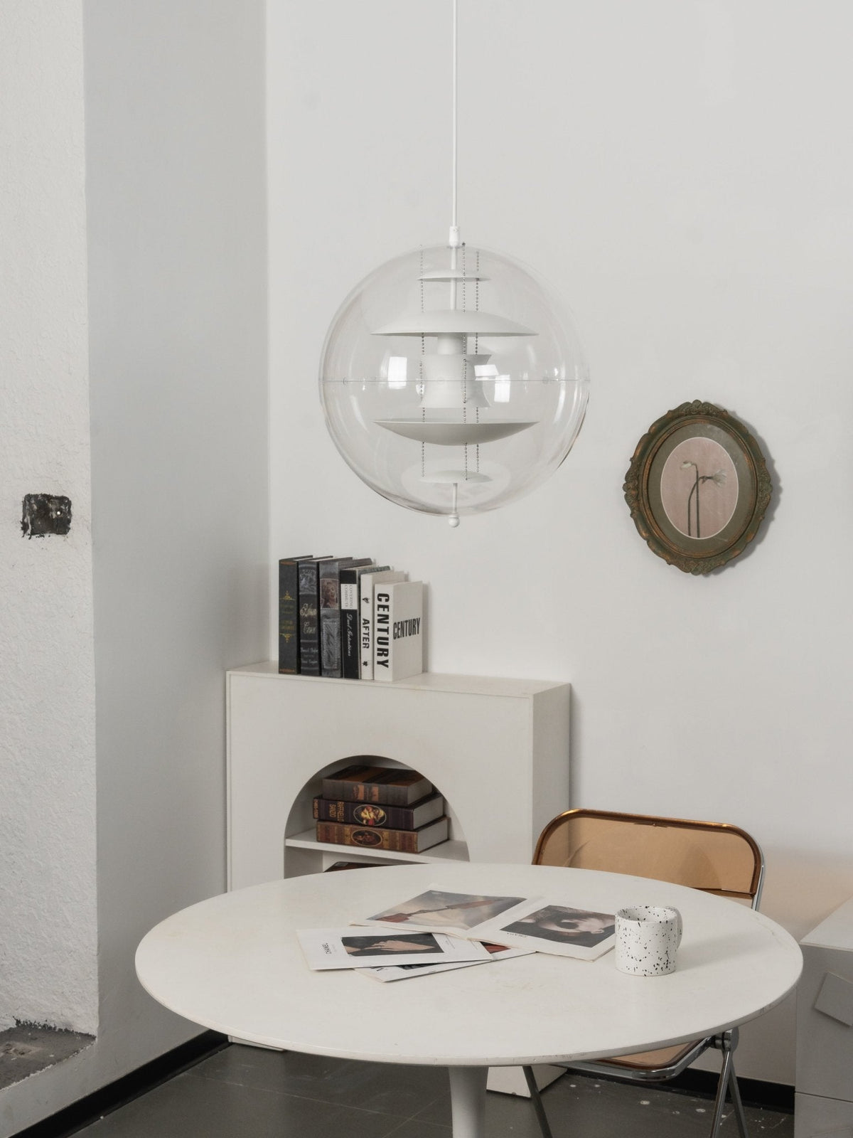 A white Clérachic sphere pendant hangs on a white cord above a round table, beside an olive-framed mirror and a book stack.