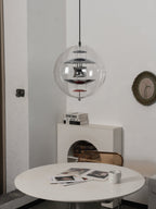 A chrome Clérachic sphere pendant, unlit, hangs above a white table with magazines, with a round olive-framed mirror on the wall.