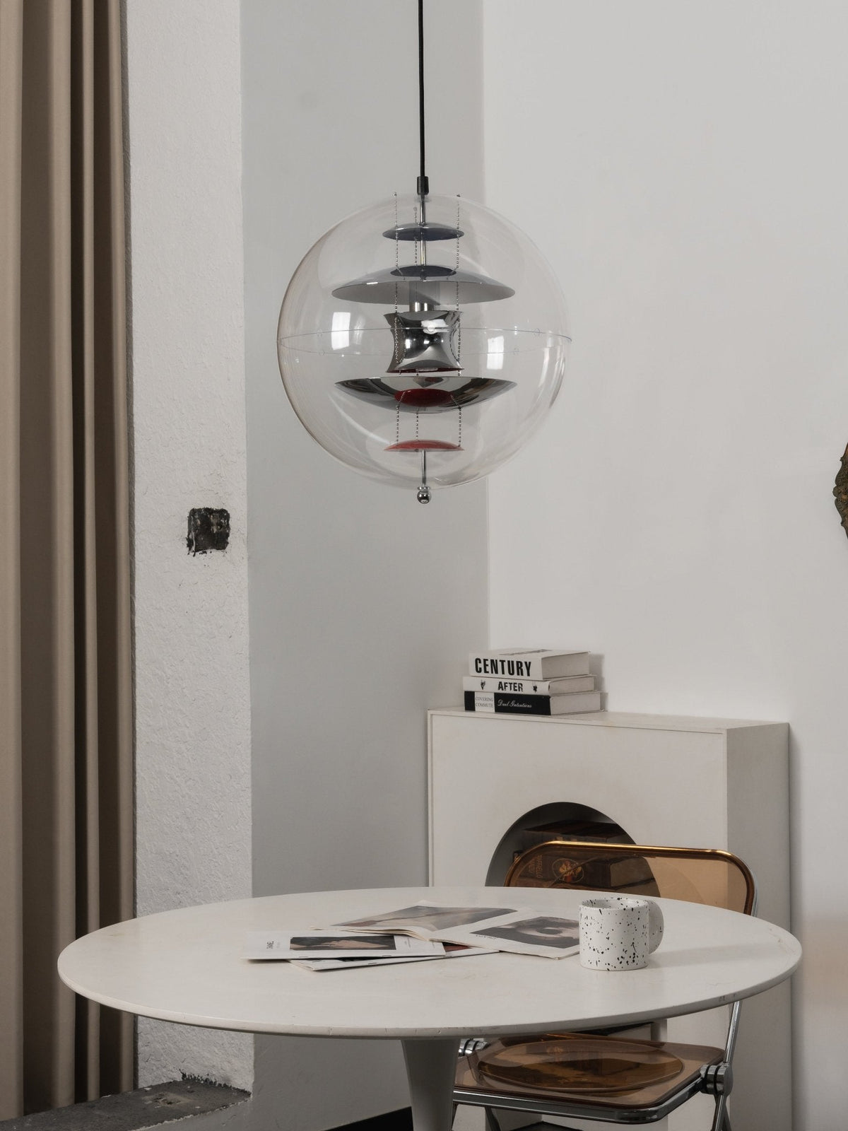 This Clérachic chrome sphere pendant hangs above a white table, with a stack of books and a clear amber chair beside it.