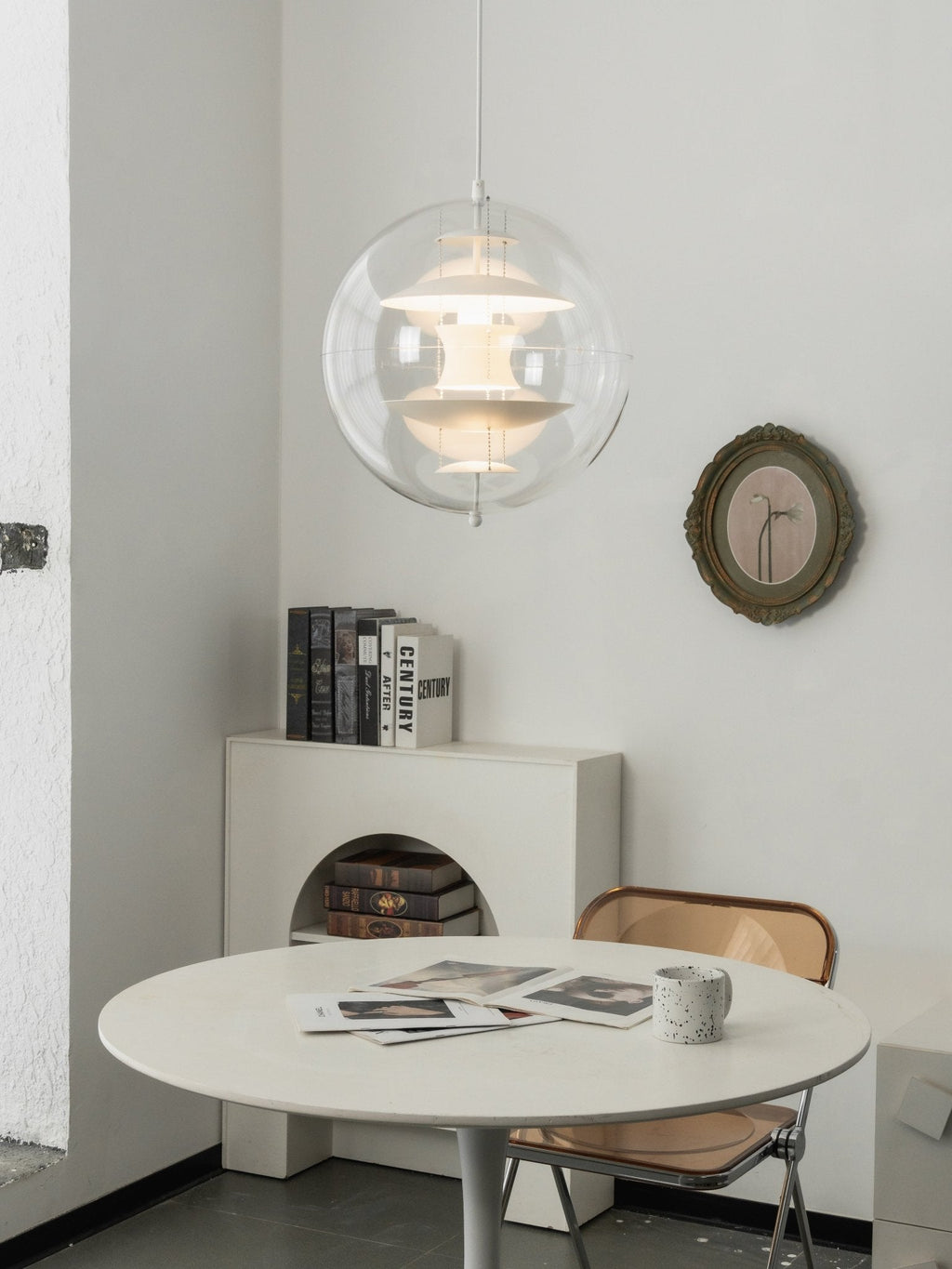 A lit white Clérachic sphere pendant glows softly above a small white round table in a corner with books and a round mirror.