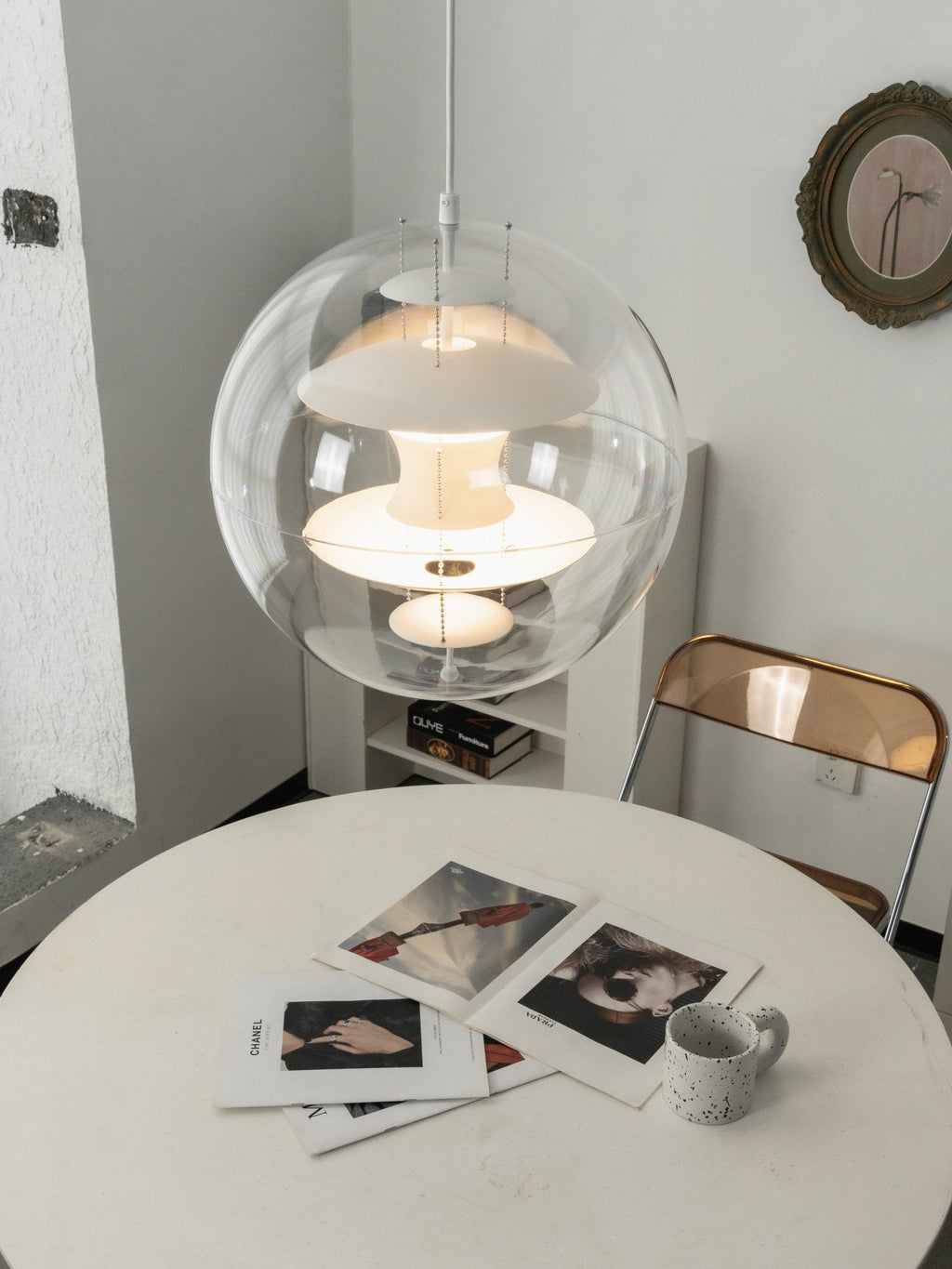 Close view of a lit white Clérachic sphere pendant hanging over a white table with printed photographs and a speckled mug.