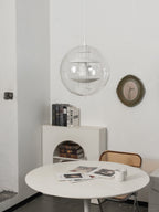 A white Clérachic sphere pendant on a white cord hangs above a white round table, with books on a shelf and a round mirror behind.