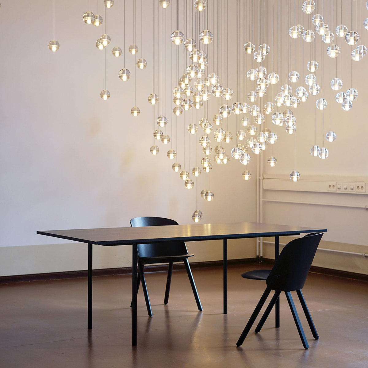 Dozens of glowing spheres from this Clérachic pendant light fill the wall of a pale room, above a long black table and two chairs.