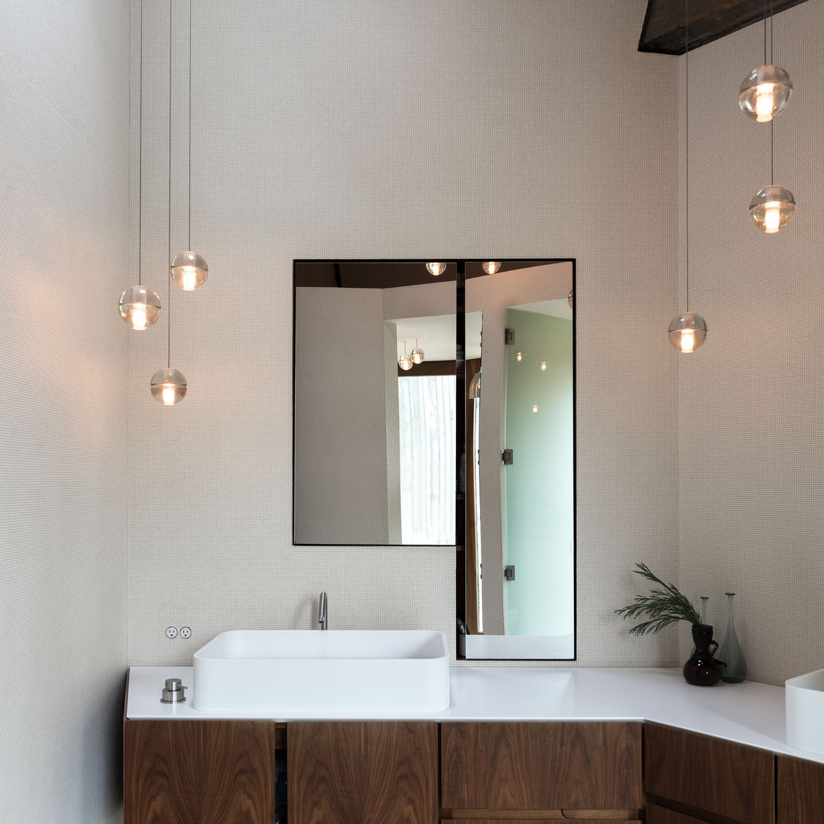 Three clear glass globes from this Clérachic pendant light hang beside a mirror above a white basin and walnut vanity.
