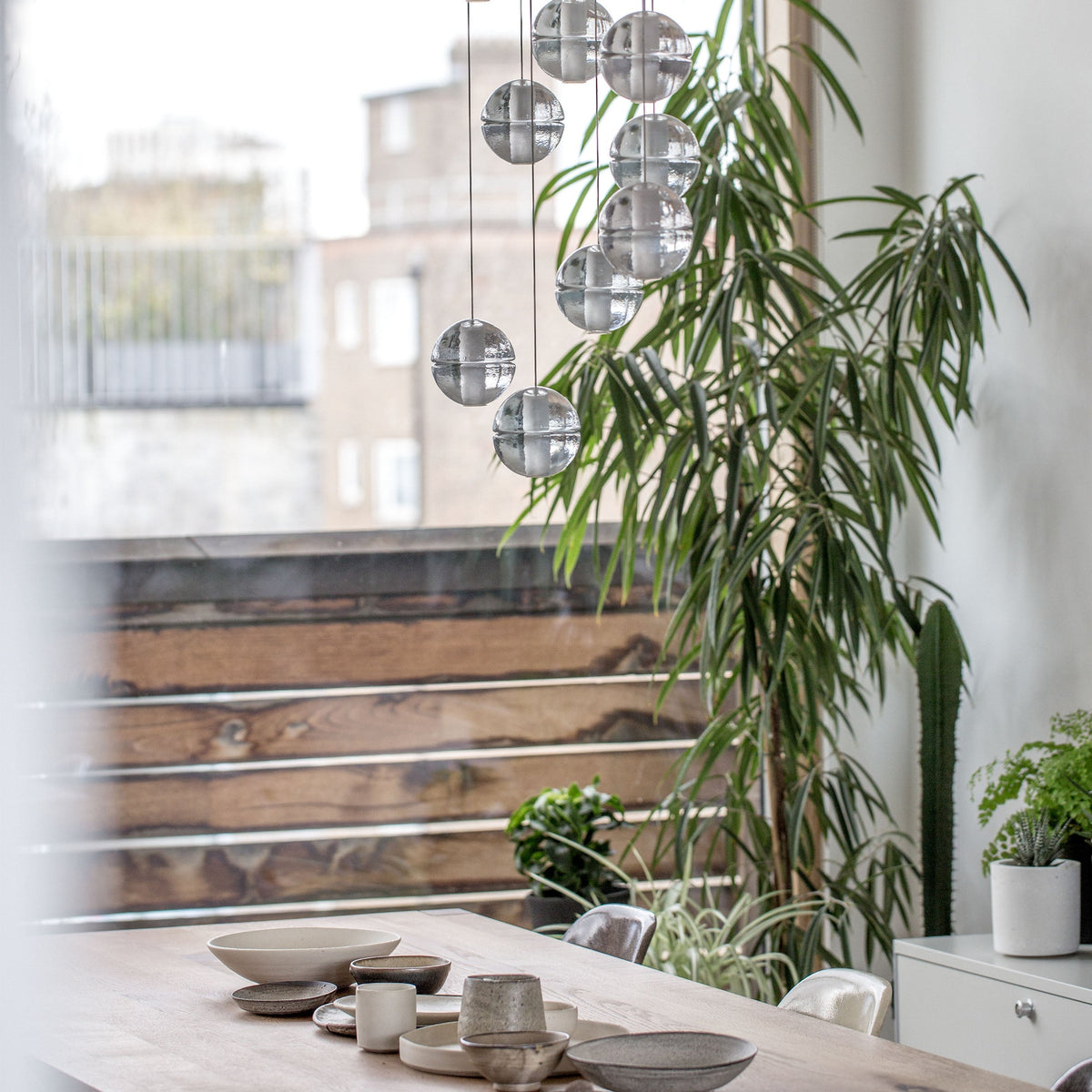 Clear bubbled glass spheres of this Clérachic pendant light hang near a terrace window, with a tall plant and a timber table beside them.