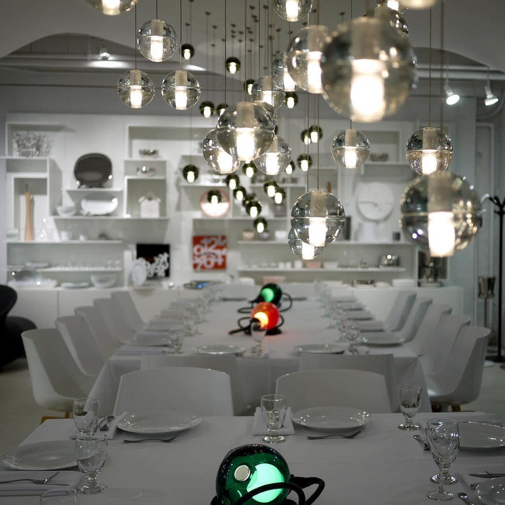 A dense group of this Clérachic pendant light's glass spheres glows above a long white-laid dining table in a bright showroom.