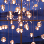 Warm lit spheres from this Clérachic pendant light hang in front of a tall window at dusk, with water and hills beyond.