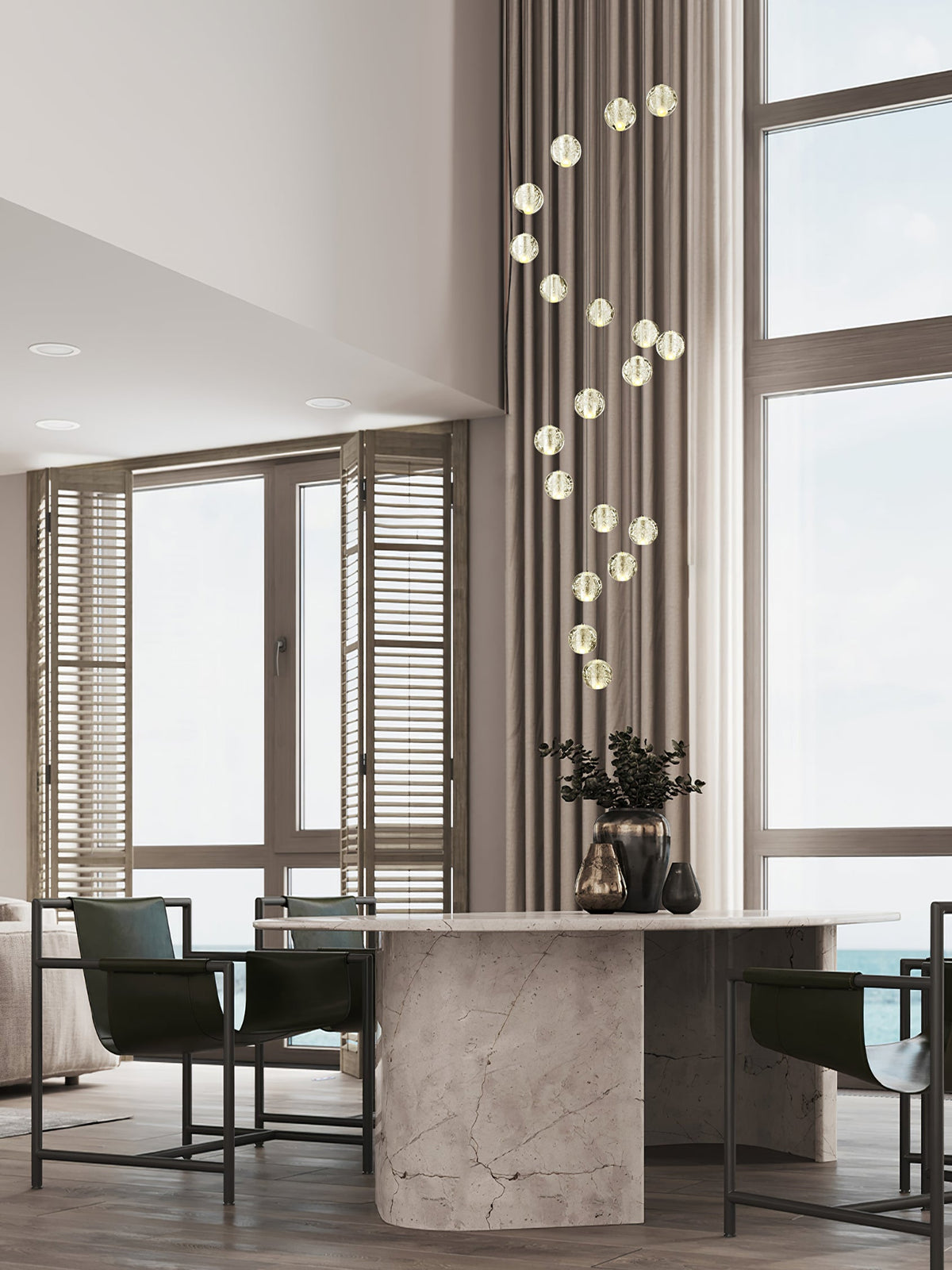 A vertical string of glowing spheres from this Clérachic pendant light rises past tall windows in a dining space with a stone table.