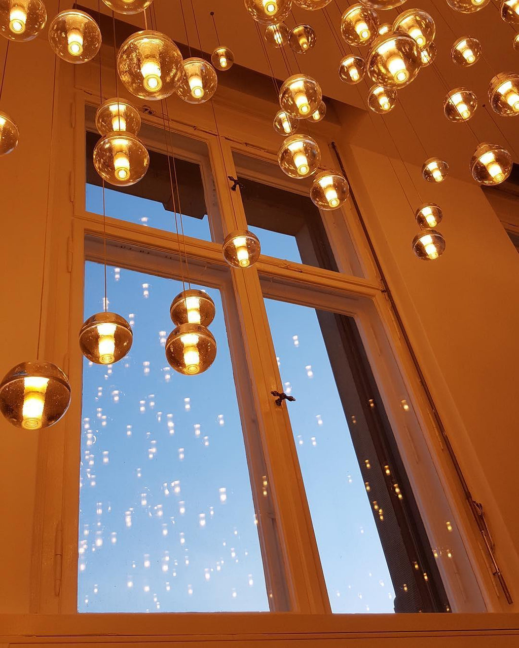 Amber-lit spheres of this Clérachic pendant light hang in front of a tall paned window, with the dusk sky showing through.