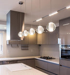 A row of four glass spheres from this Clérachic pendant light hangs at different heights in a grey kitchen under a steel hood.