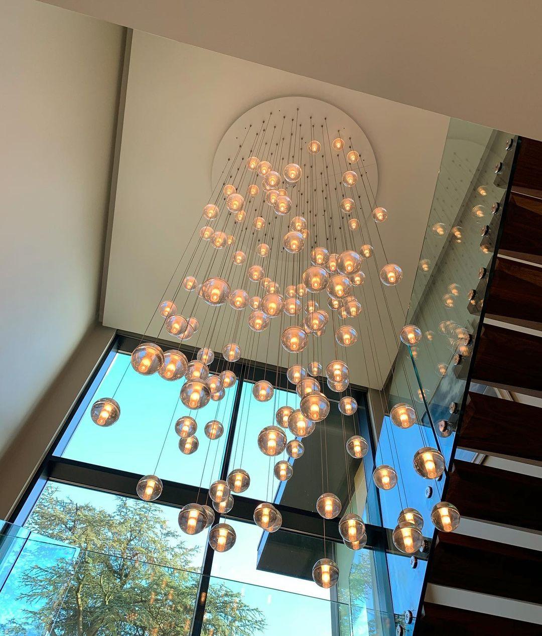 A tall cascade of warm lit spheres from this Clérachic pendant light falls from a round ceiling plate beside a glass stair.