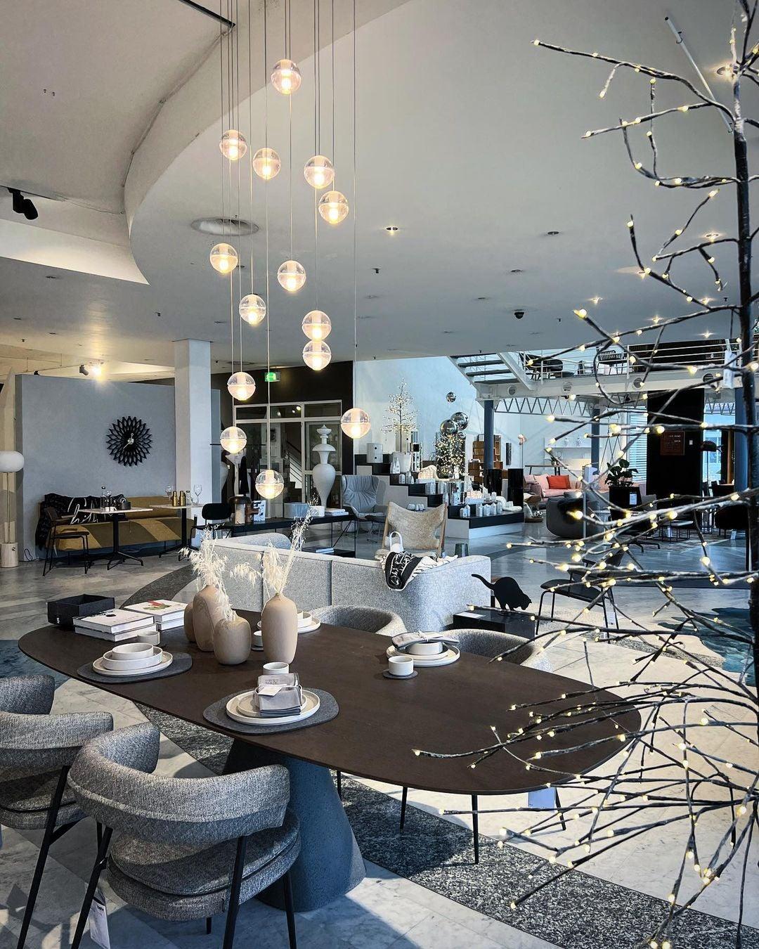 Spheres of this Clérachic pendant light hang over a dark oval table in a large open-plan showroom with grey chairs.