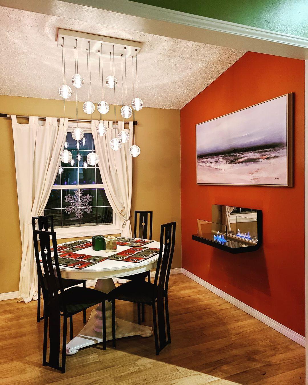 Over a round dining table in a room with an orange wall, this Clérachic pendant light hangs spheres from a long rectangular canopy.