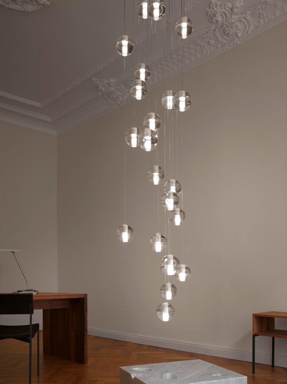 A column of lit glass spheres from this Clérachic pendant light drops through a tall, ornate-ceilinged room to a wooden desk below.