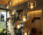 Large warm-lit spheres of this Clérachic pendant light hang close to the camera, with a window and plants blurred behind.
