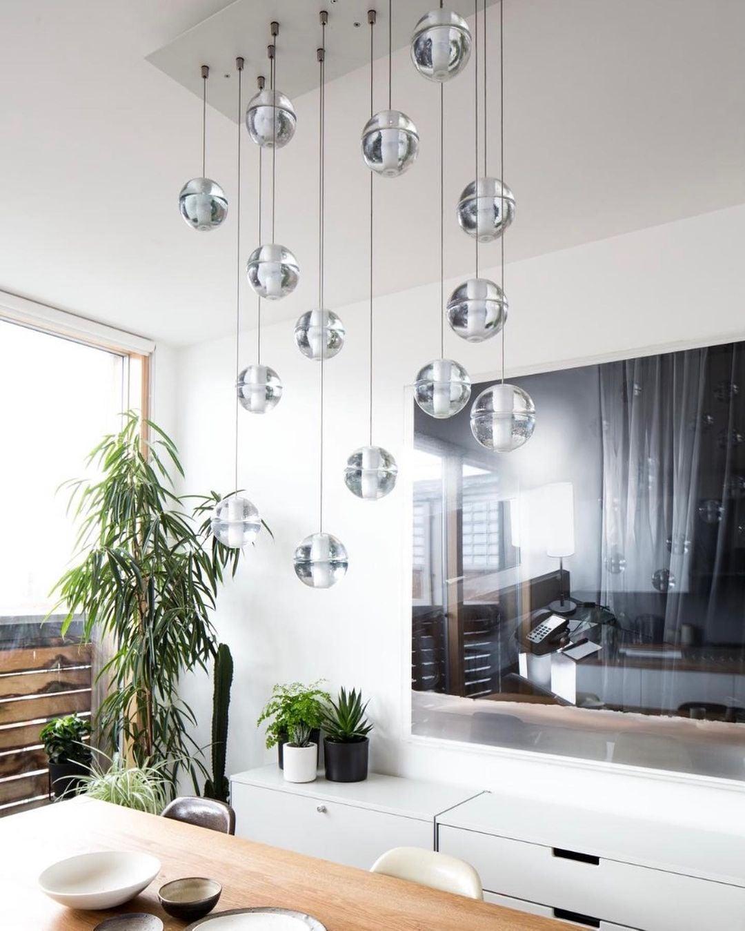A grid of clear glass spheres from this Clérachic pendant light hangs from a flat white ceiling plate in a bright room with a tall plant.