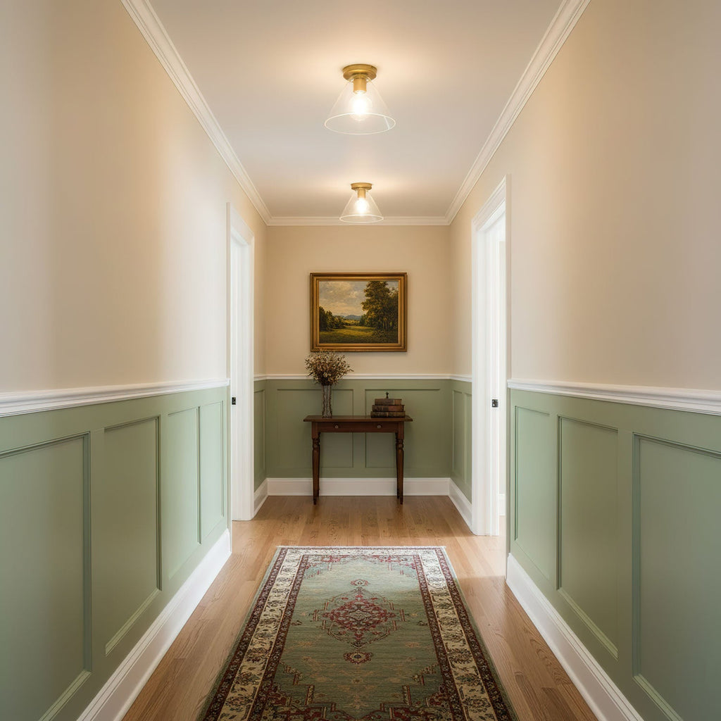 Two brass ceiling lights with clear glass shades glow down a hallway with sage-green panelling and a patterned runner, from Clérachic.