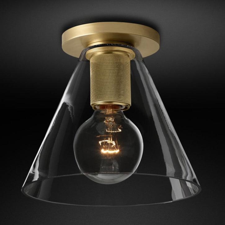 Close view of the brass base and knurled socket, with a filament bulb inside a clear conical glass shade, on a black backdrop, from Clérachic.