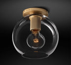 Round clear glass globe around a bare filament bulb, with a brass base and knurled socket on a dark background, from Clérachic.