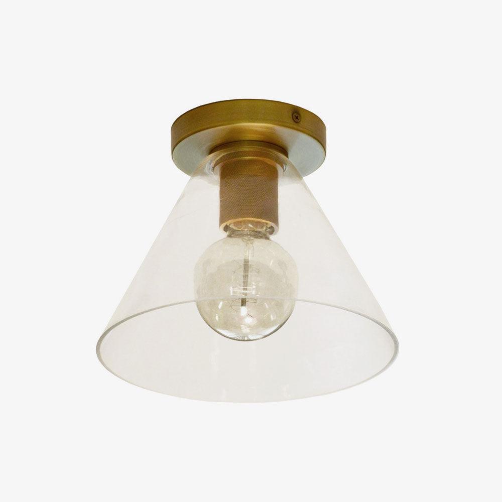 Brass-based flush light with a clear cone glass shade and a round bulb inside, on a pale backdrop, from Clérachic.