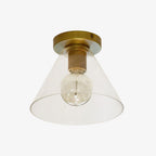 Brass-based flush light with a clear cone glass shade and a round bulb inside, on a pale backdrop, from Clérachic.