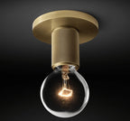 Close view of the knurled brass socket and a glowing filament bulb below the round brass plate, on this Clérachic flush mount.