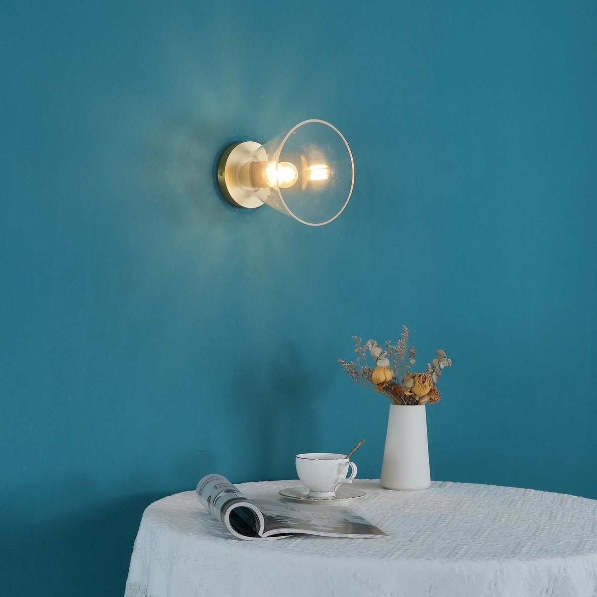 Brass-backed light with a clear glass shade fixed to a teal wall, lit, with a white vase of dried flowers and a cup below, from Clérachic.