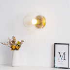 Clear round glass shade and brass fitting mounted on a white wall above a vase of dried flowers and a framed print, from Clérachic.