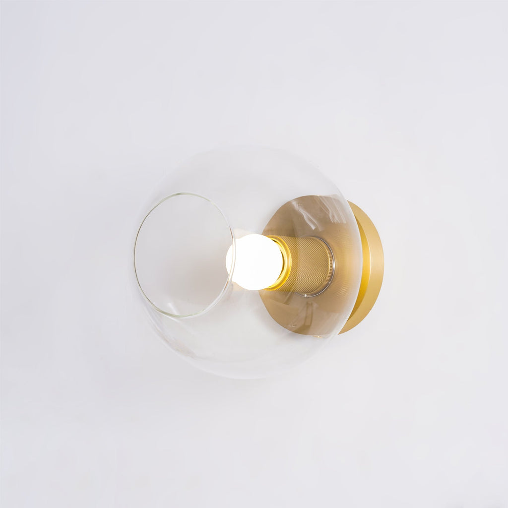 Side-on view of the clear glass globe, brass socket and lit white bulb mounted on a pale wall, from Clérachic.