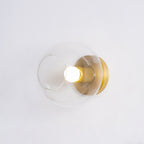 Side-on view of the clear glass globe, brass socket and lit white bulb mounted on a pale wall, from Clérachic.