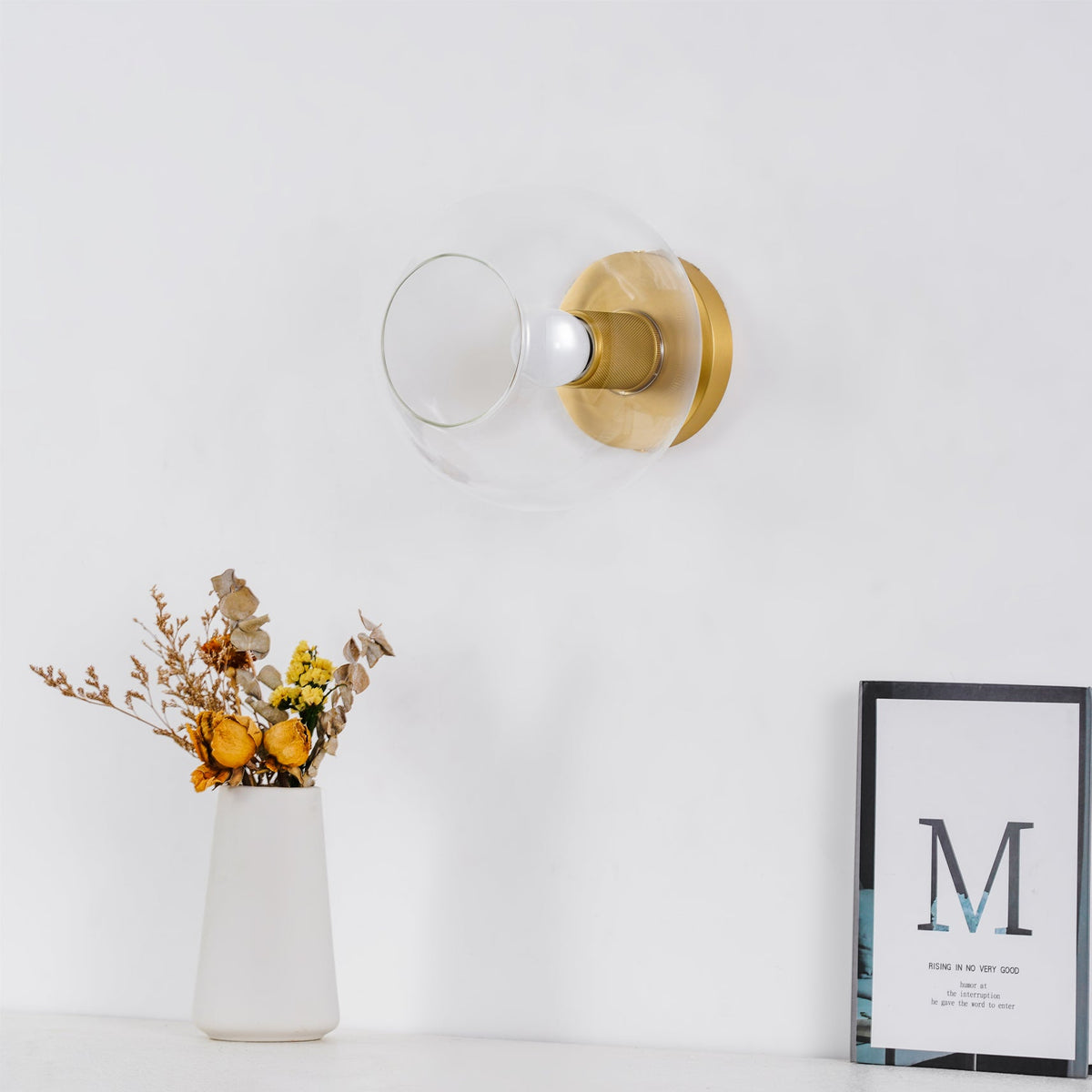 Small globe-shaped light on a white wall, lit, above a white vase of dried flowers and a framed print, a Clérachic flush mount.