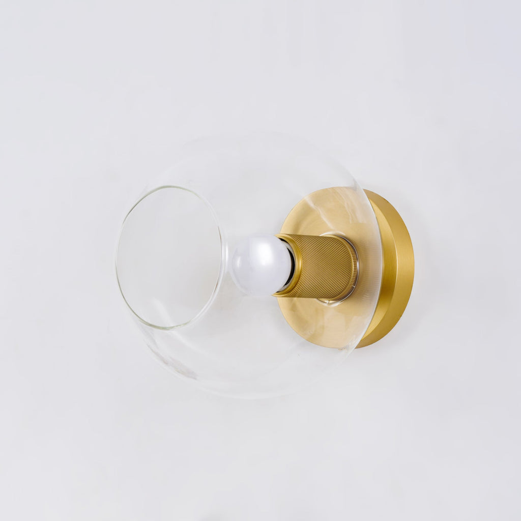 Close view of the clear glass globe with a brass backplate and a knurled brass socket holding a lit bulb, from Clérachic.