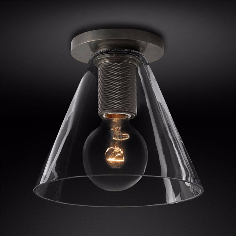 Black metal plate and knurled socket holding a filament bulb inside a clear conical glass shade, on a dark background, from Clérachic.