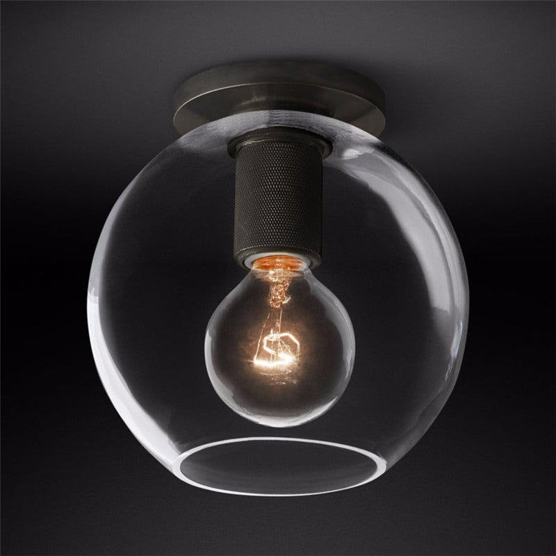 Clear glass globe around a glowing bulb, with a black plate and knurled black socket, against a dark backdrop, a Clérachic flush mount.