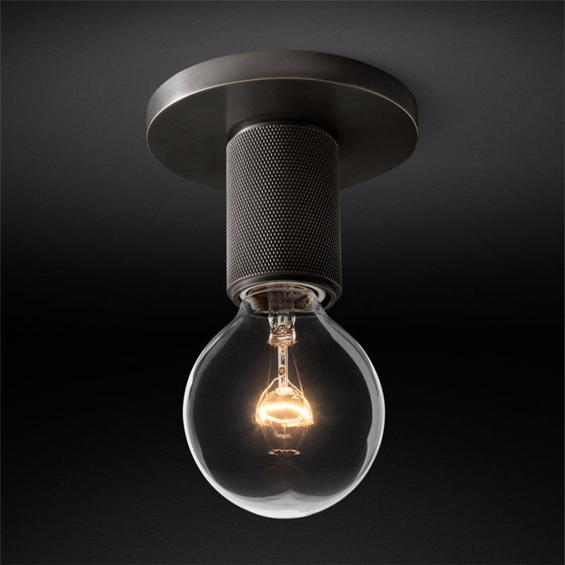 Close view of a black ceiling plate, knurled socket and bare globe bulb lit warm, on a dark ground, from Clérachic.