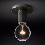 Close view of a black ceiling plate, knurled socket and bare globe bulb lit warm, on a dark ground, from Clérachic.