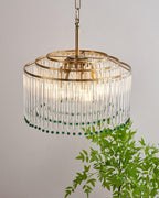 Seen from slightly below, this Clérachic chandelier shows three gold rings, hanging glass rods and green beads, beside a leafy branch.