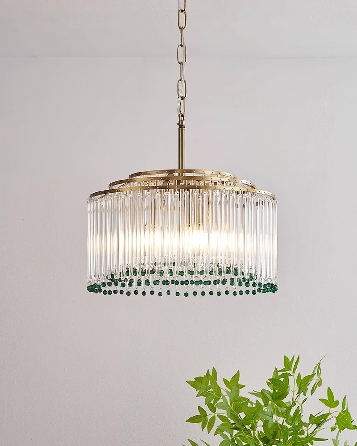 A Clérachic gold and glass rod chandelier on a straight stem and chain, with a leafy plant in the corner of the frame.