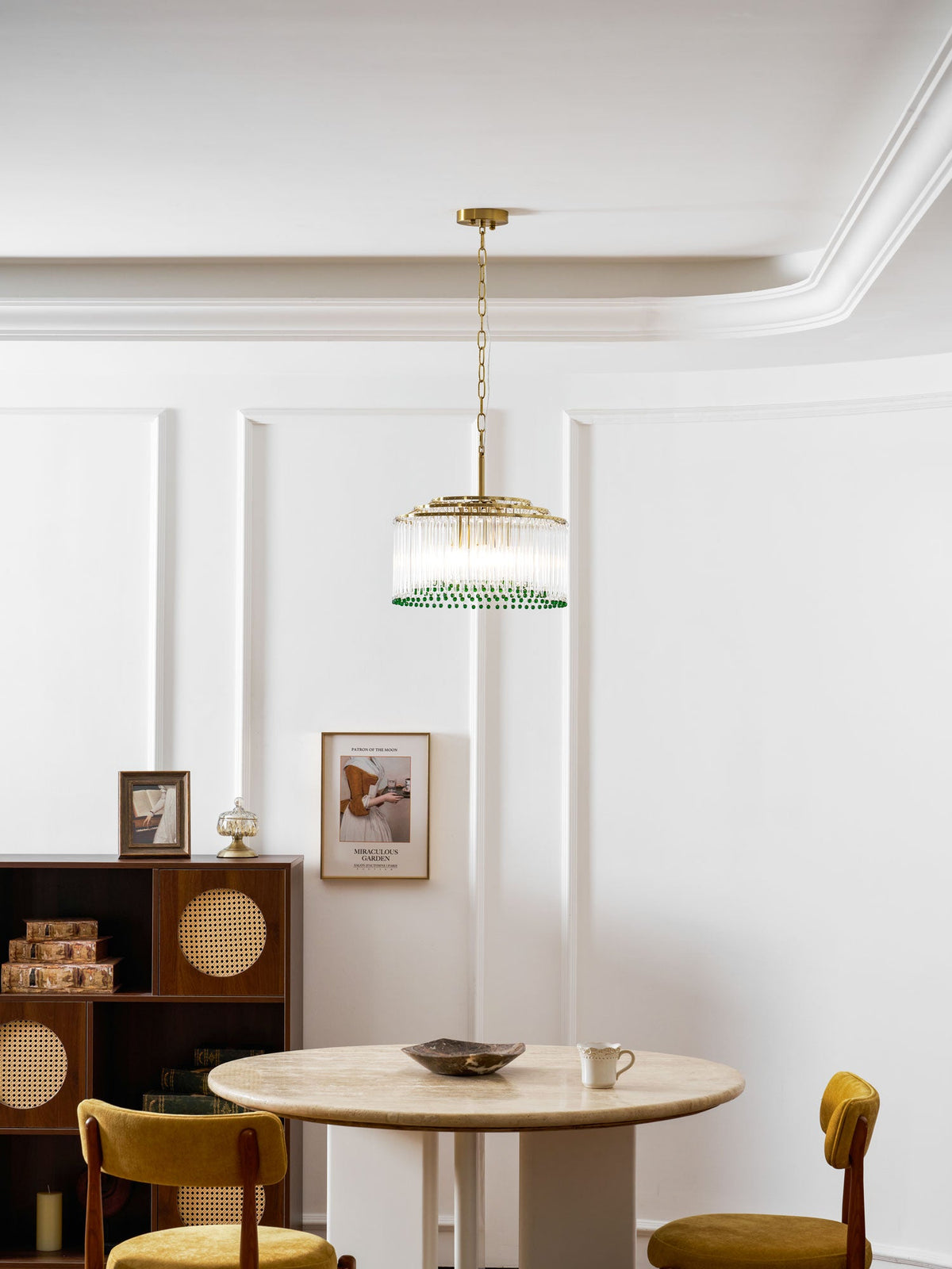 A Clérachic green-beaded chandelier hangs over a round table with a shallow dish, mustard chairs and a wooden shelf unit.