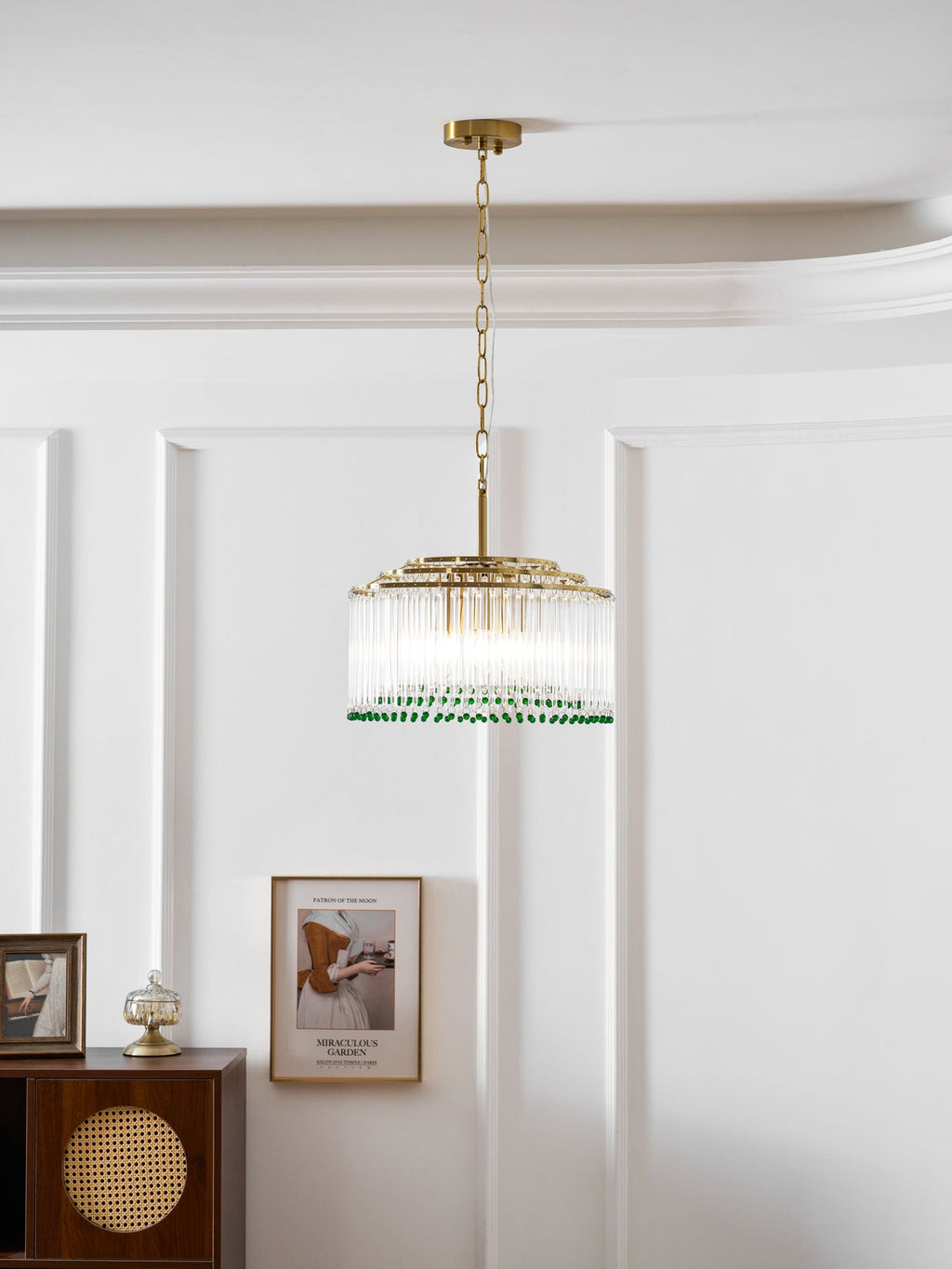 A Clérachic glass rod chandelier with a gold chain hangs on a white panelled wall beside a framed print and a cabinet.