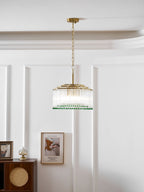 A Clérachic glass rod chandelier with a gold chain hangs on a white panelled wall beside a framed print and a cabinet.