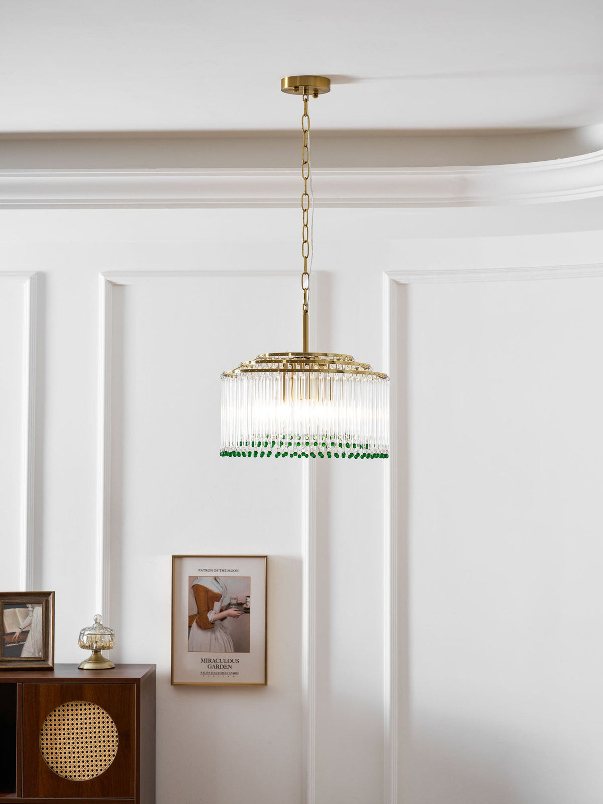 A Clérachic glass rod chandelier with a gold chain hangs on a white panelled wall beside a framed print and a cabinet.