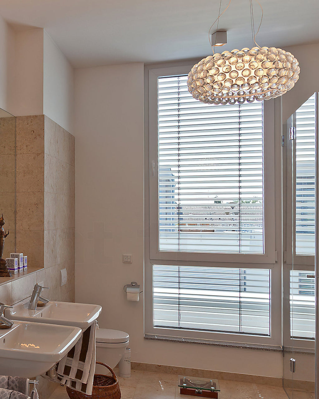A clear sphere pendant glows warm in front of a tall shuttered window in a pale bathroom with twin basins, from Clérachic.